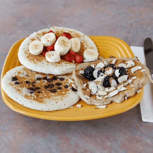 Three pancakes on a yellow plate, topped with bananas, strawberries, chocolate chips, blackberries, and almonds.
