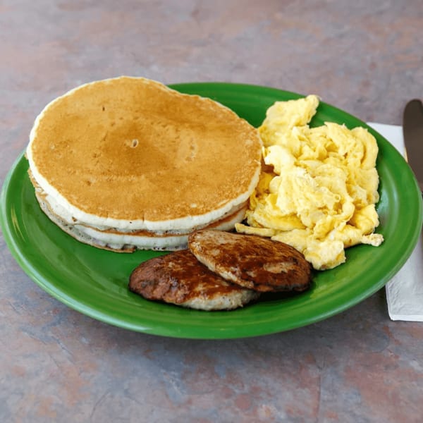 Breakfast plate with a stack of pancakes, scrambled eggs, and two sausage patties.