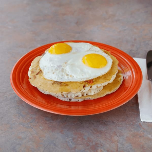 Two fried eggs with bright yellow yolks on a stack of pancakes on an orange plate.