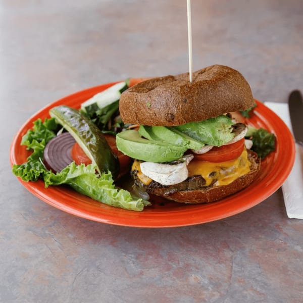 A burger with avocado, cheese, mushrooms, and tomato, served with a pickle and salad.